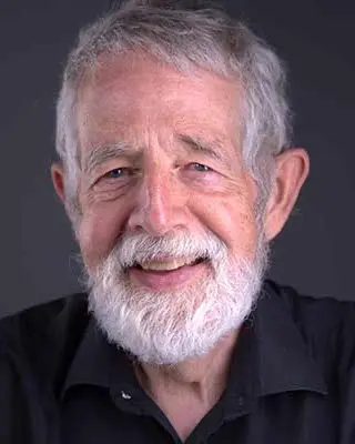 Older man with gray hair and beard, wearing a black collared shirt, smiling at the camera against a plain dark background.
